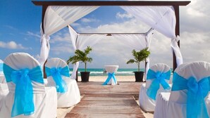 Un muelle de madera que conduce a un montaje para boda en la playa con sillas blancas y cintas azules, y dos palmeras en macetas.