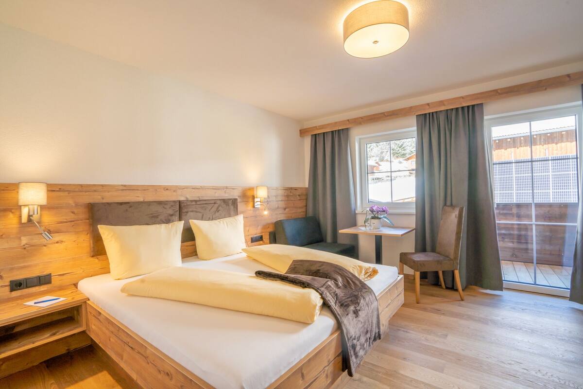 A hotel room with a wooden headboard, a large bed with white linens, a bedside table with a lamp, a small table with a chair, and a window with curtains.