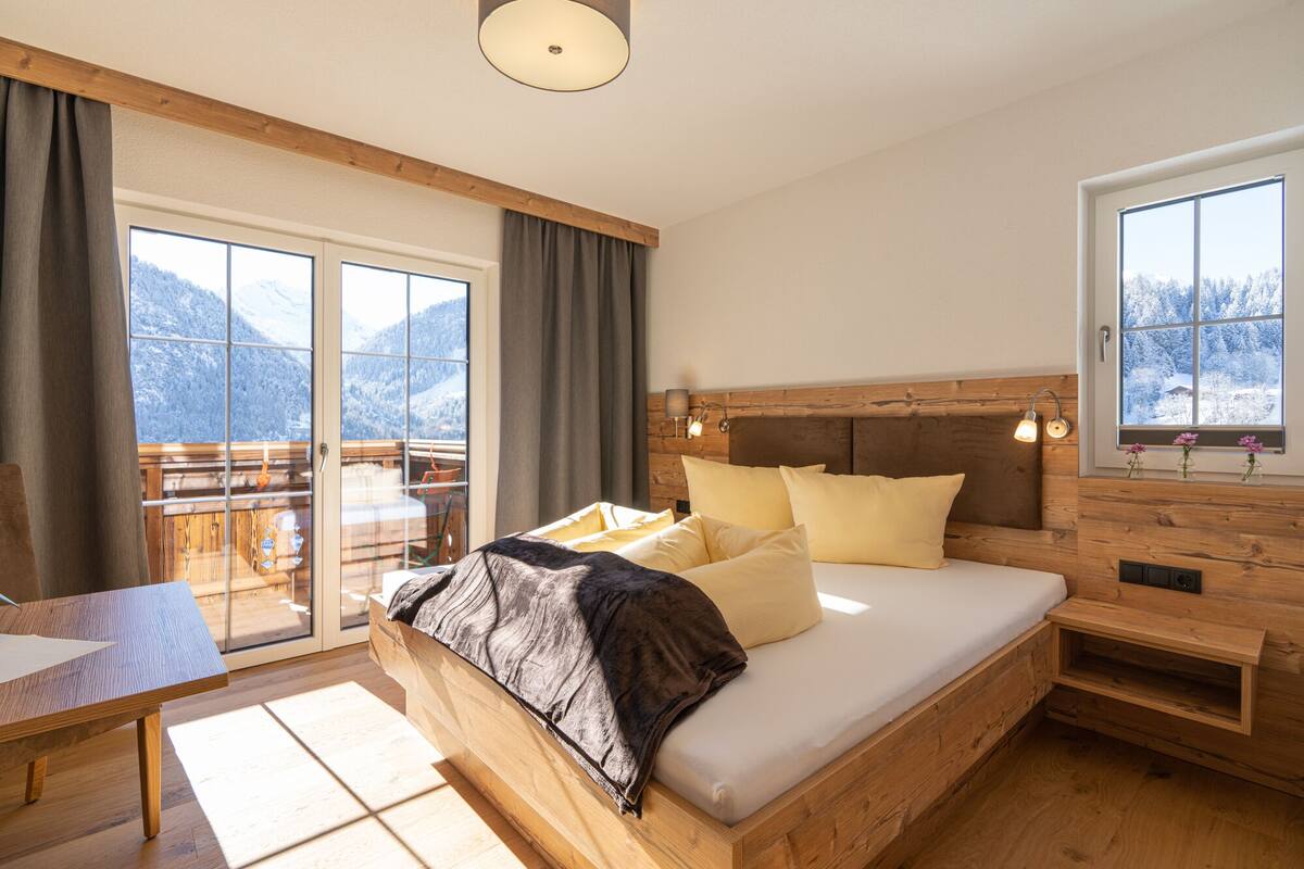 A bedroom with a wooden bed, a wooden headboard, a nightstand, a desk, and a window with a view of snow-covered mountains.