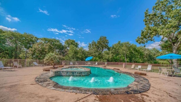 A circular pool with a hot tub, surrounded by lounge chairs and umbrellas, with trees in the background.