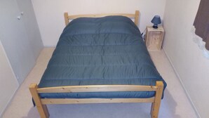 A small, single-bed room with a wooden frame, a blue mattress, and a small wooden nightstand with a lamp.