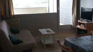 A living room with a sofa, a small table, a TV on a stand, and a window with blinds.