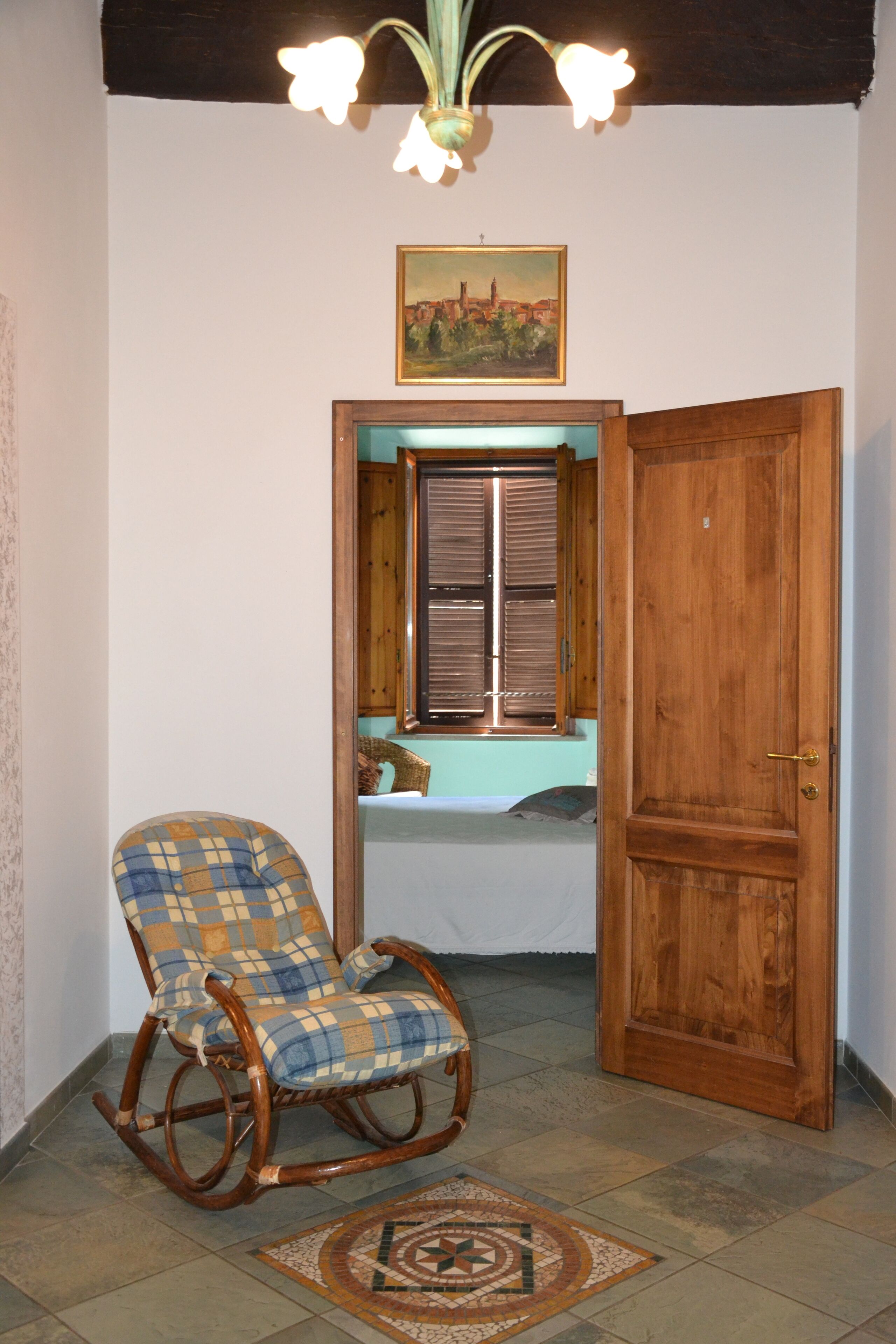 Een kamer met een schommelstoel, een houten deur, een raam met luiken en een hanglamp.