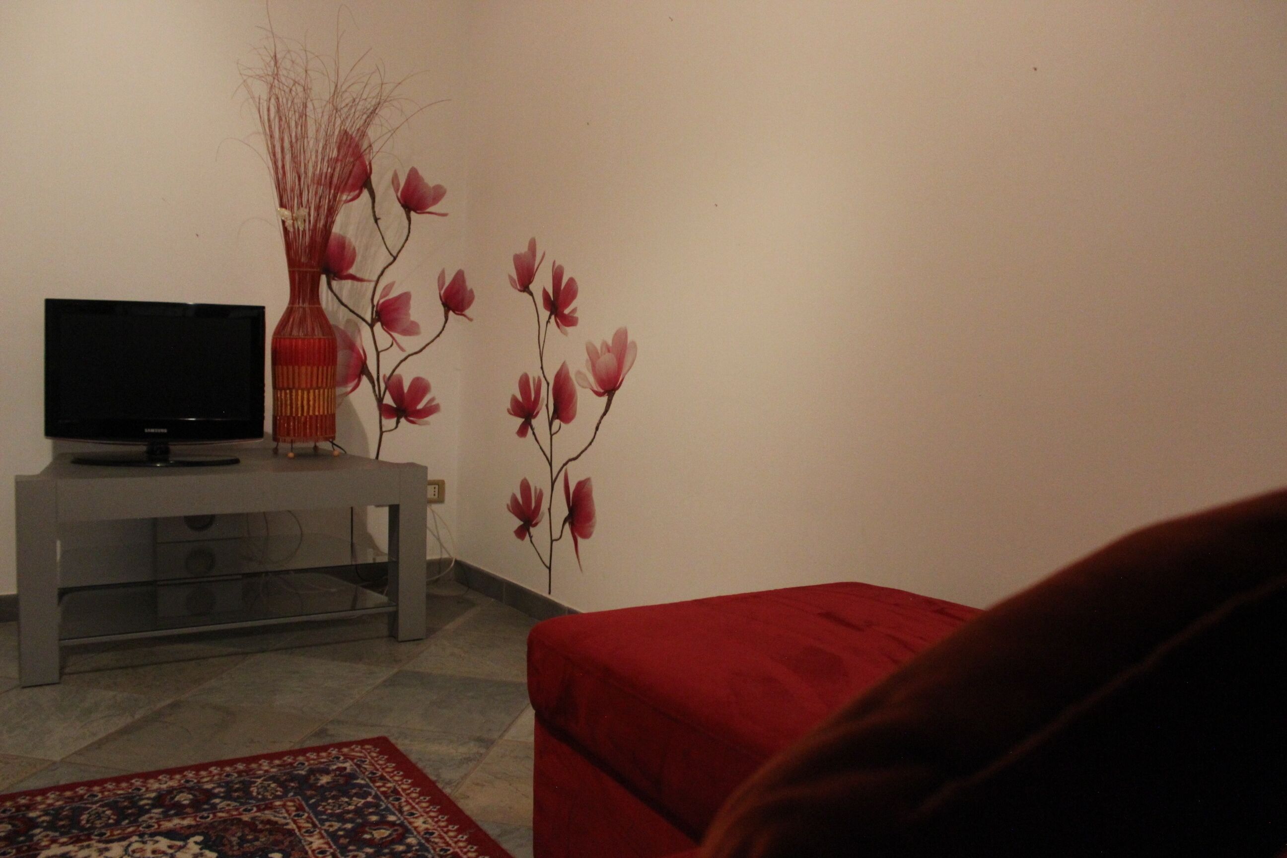 Una sala de estar con un televisor sobre un mueble gris, un sofá rojo y un jarrón decorativo con ramas y flores en una pared blanca.