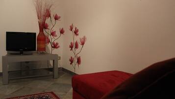 Una sala de estar con un televisor sobre un mueble gris, un sofá rojo y un jarrón decorativo con ramas y flores en una pared blanca.