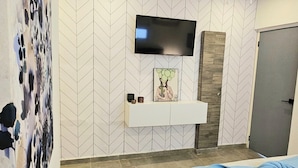 Une chambre moderne avec un téléviseur à écran plat fixé au mur, un lit recouvert d’une couverture bleue et un tapis à motifs sur le sol.