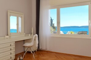 A room with a wooden floor, a white dresser with drawers, a chair, a window with a view of the sea, and a curtain.