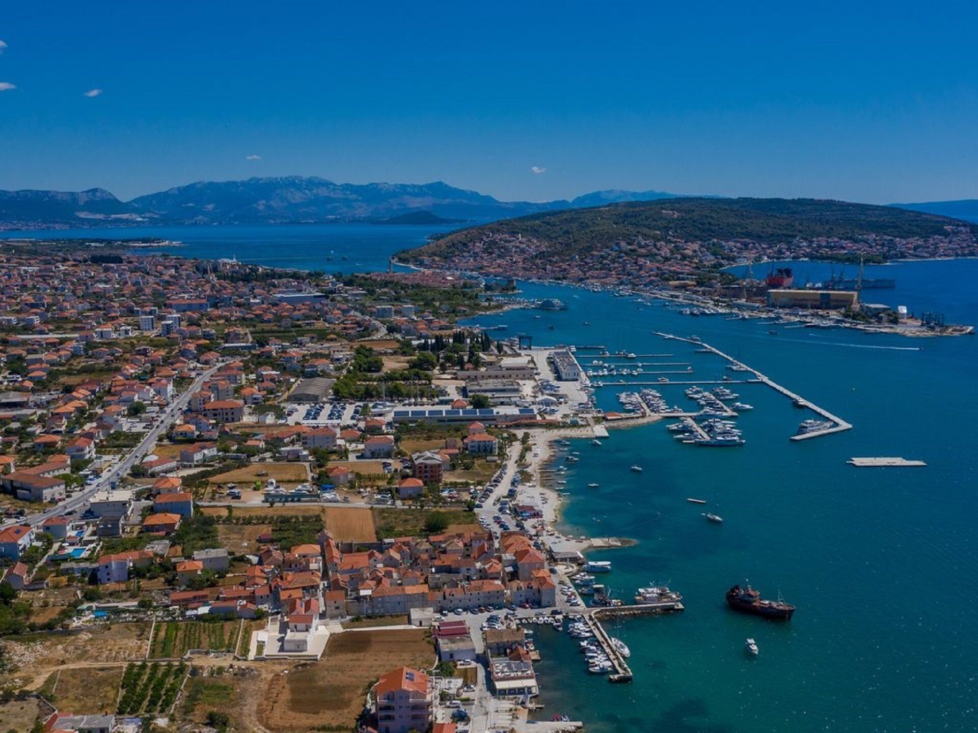 Vista aérea de un pueblo costero con un puerto deportivo, zonas residenciales y montañas al fondo.