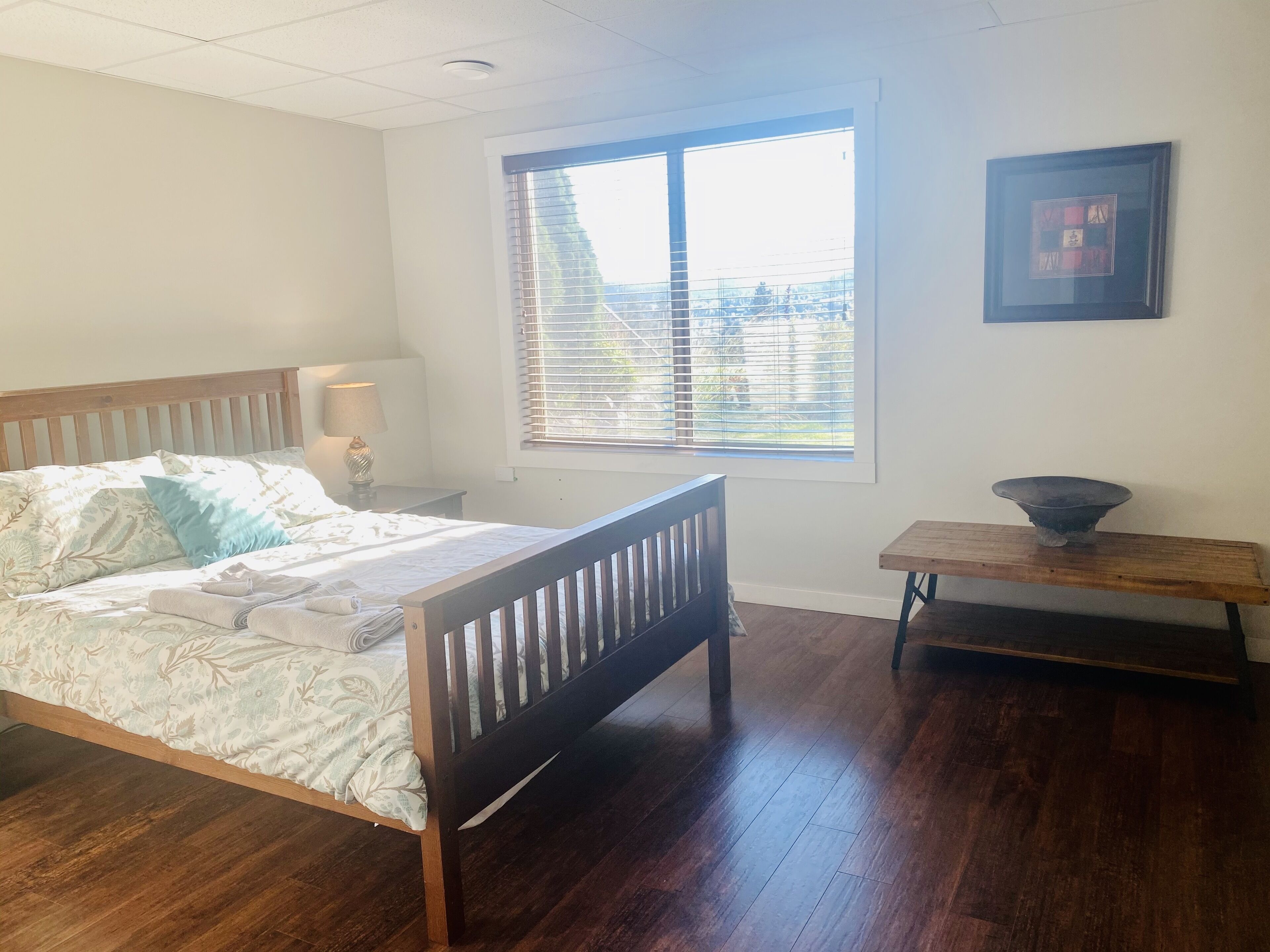 A bedroom with a wooden bed, a bedside table with a lamp, a window with blinds, and a framed picture on the wall.