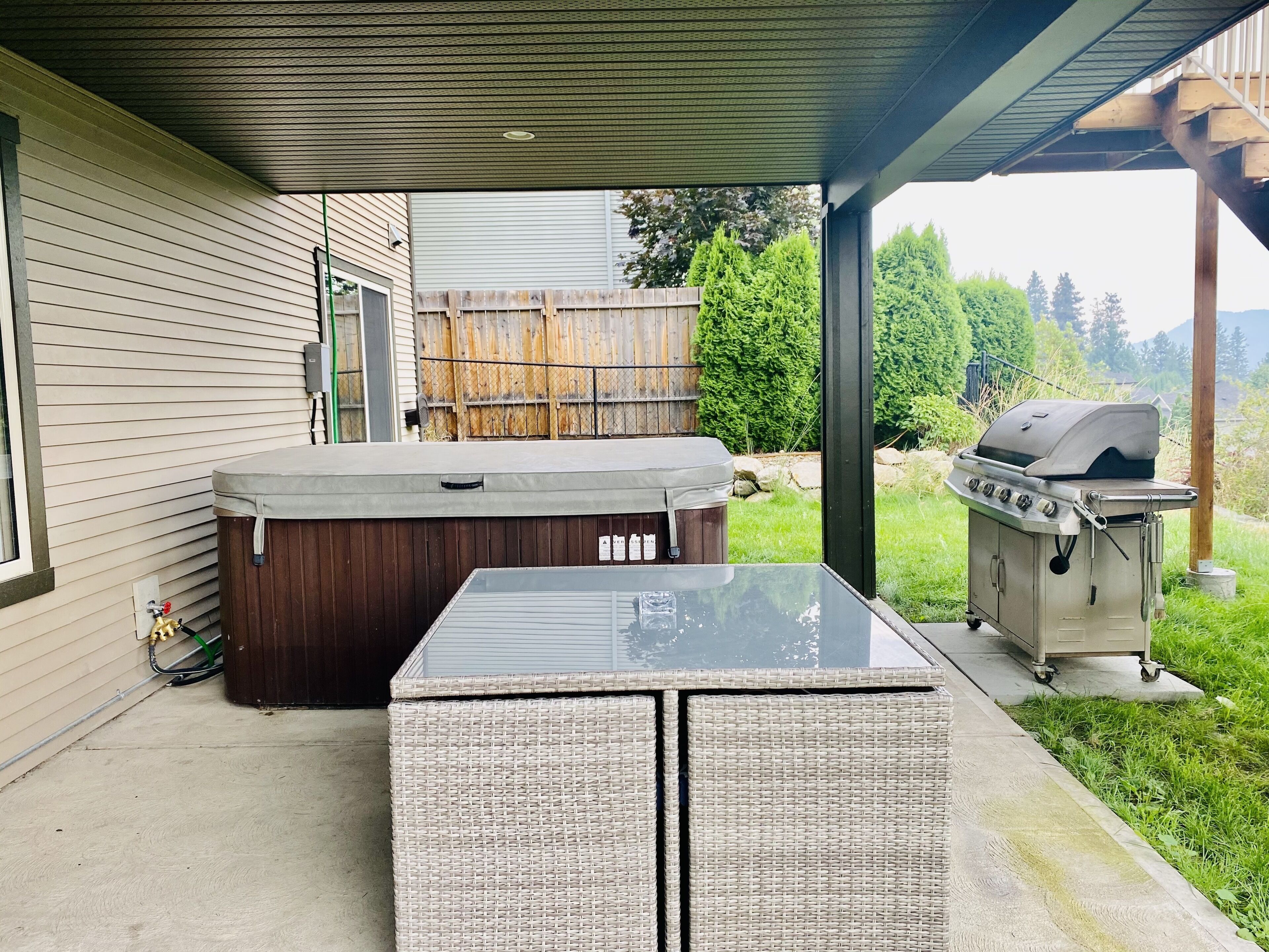 A covered patio with a hot tub, grill, and wicker furniture.