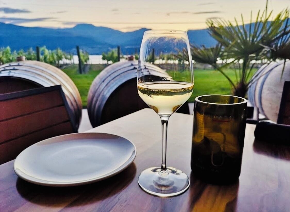Uma mesa com uma taça de vinho e uma caneca, um prato e dois barris ao fundo.