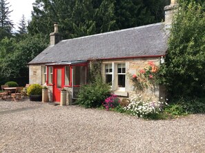 Une charmante maison en pierre avec une porte rouge, une allée gravillonnée et une petite terrasse aménagée avec du mobilier d’extérieur.