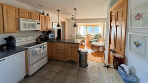 A kitchen with wooden cabinets, a microwave, stove, and a trash can. There is a dining area with chairs and a table visible through an open door.