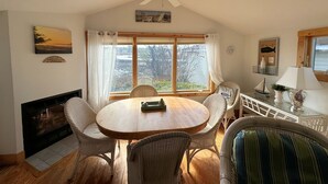 A cozy dining area with a round wooden table surrounded by wicker chairs, a fireplace, and a large window with curtains.