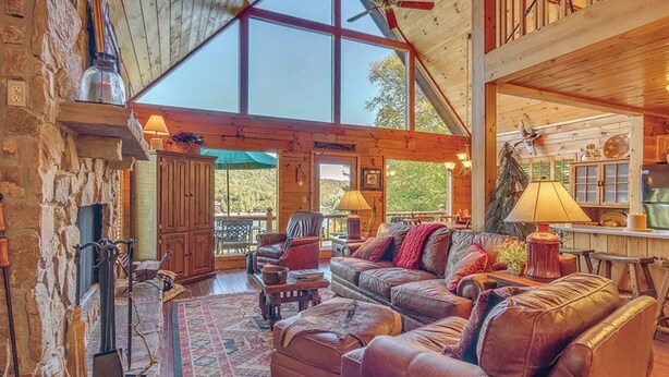 Ein gemütliches Blockhaus-Interieur mit Kamin aus Stein, Ledersofas und einem großen Fenster mit Blick ins Freie.