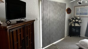 Une pièce avec un téléviseur posé sur un meuble en bois, une porte coulissante, un ventilateur de plafond, un tableau encadré et une petite table sur laquelle est disposé un arrangement floral.