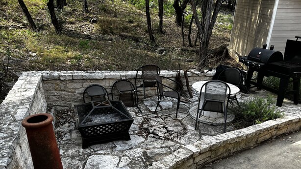 Un foyer en pierre, agrémenté de chaises et d’une table, entouré d’un mur de pierre et de végétation.