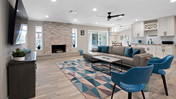 Una sala de estar moderna con sofá, mesa de centro y sillas azules. Se observa una chimenea, un televisor y una cocina al fondo.