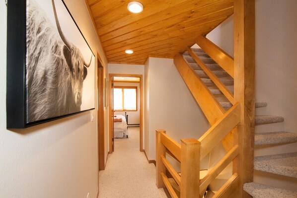 Een hal met een houten trap, een ingelijst schilderij aan de muur, en een deur die naar een kamer leidt.