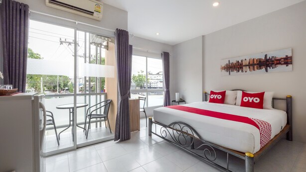 A hotel room with a bed, a balcony with chairs, a cityscape painting, and a view of buildings outside. - OYO 75323 Tha Burapa Home Bangsean (Chonburi)