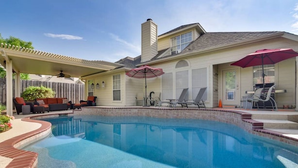 Une piscine avec un patio couvert, des meubles d’extérieur et un parasol rouge.