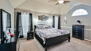 Une chambre à coucher comprenant un lit, une commode, une table de chevet, un ventilateur de plafond et une fenêtre.