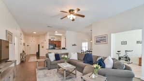 A spacious living room with a grey sofa, coffee table, and a ceiling fan. A kitchen is visible in the background with a dining area.