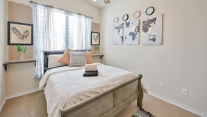 A bedroom with a bed, a ceiling fan, a window with curtains, and wall art including clocks and maps.
