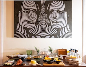 A dining table with food and drinks, a painting above it, and a window with natural light. - Hotel Rural Quinta do Juncal (Peniche)