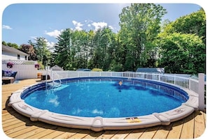 A round above-ground pool with a wooden deck, surrounded by a white fence, and trees in the background.