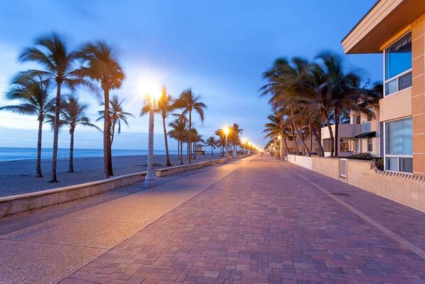Stroll along the beachfront promenade lined with palm trees and paved with red bricks, leading to a modern hotel with a pool area overlooking the ocean.