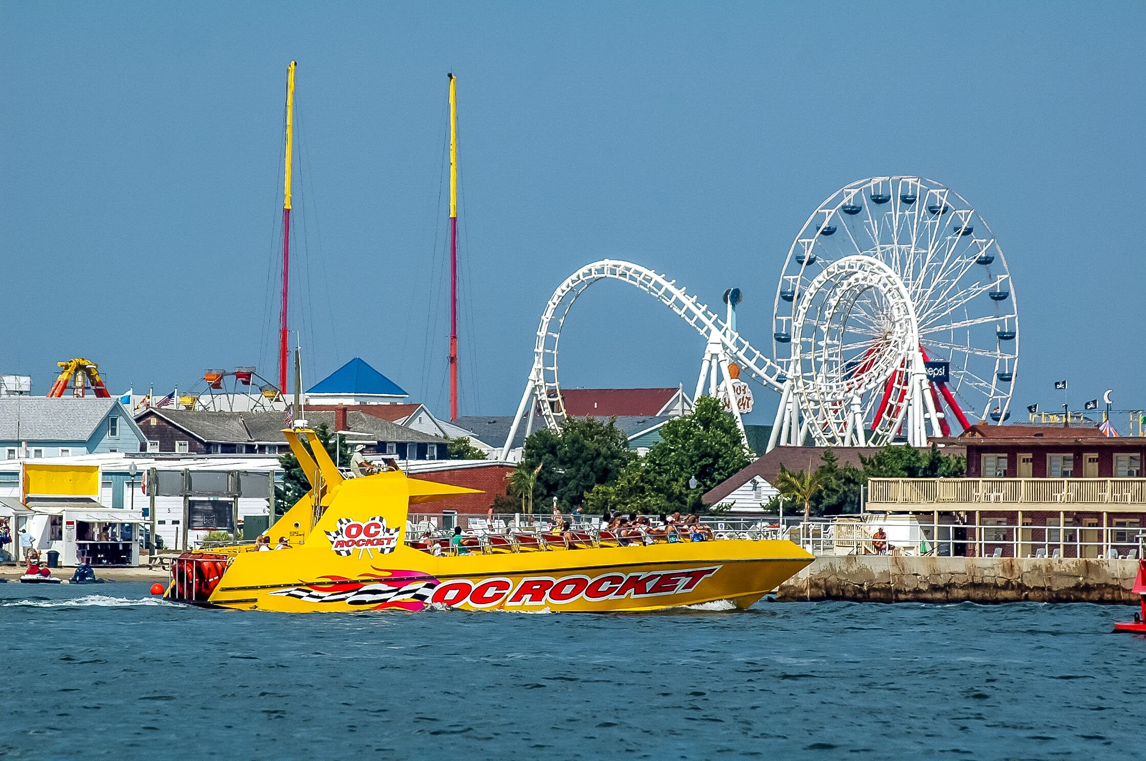 A yellow speedboat with 'DC ROCKET' branding on the side, docked near a waterfront with a Ferris wheel and other structures in the background.