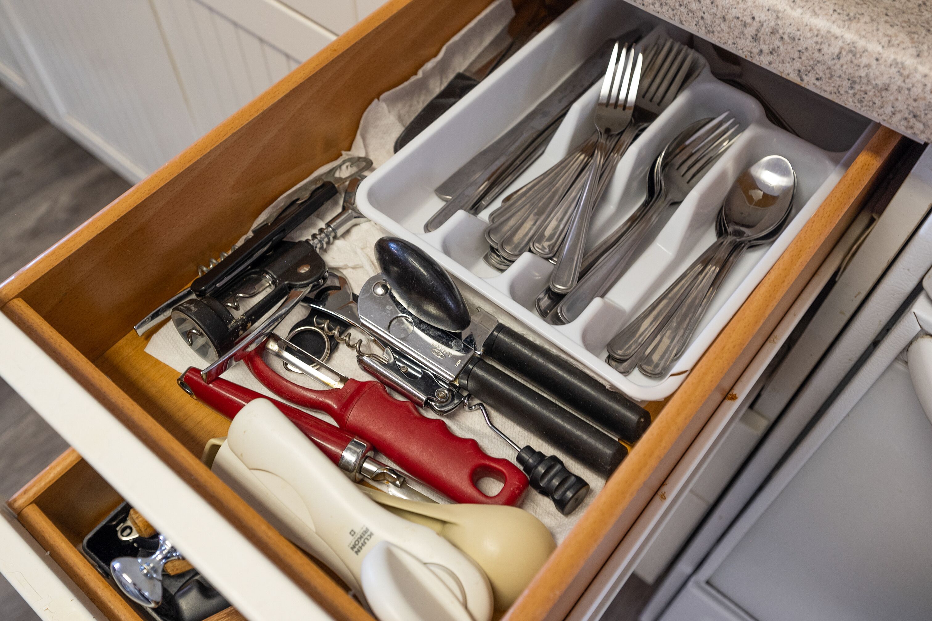 A drawer containing various kitchen utensils including a red can opener, a white can opener, a black can opener, a silver spoon, a silver fork, and a silver knife.