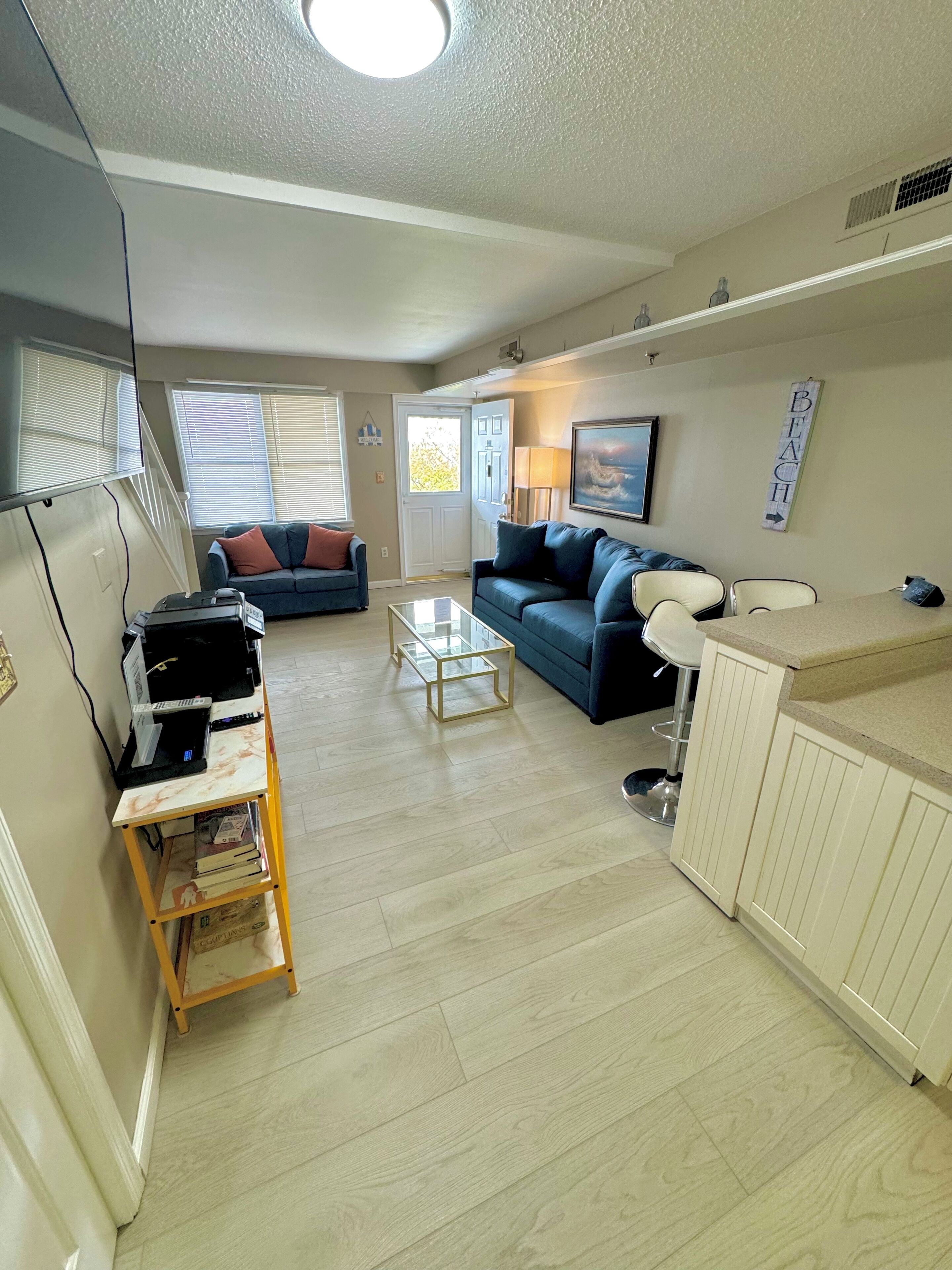 A living room with a sofa, coffee table, and a small bookshelf. There is a door leading to another room and a window with blinds.