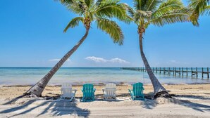 Une plage avec trois chaises sous des palmiers, une jetée en bois qui s’avance dans l’eau et un ciel bleu clair.