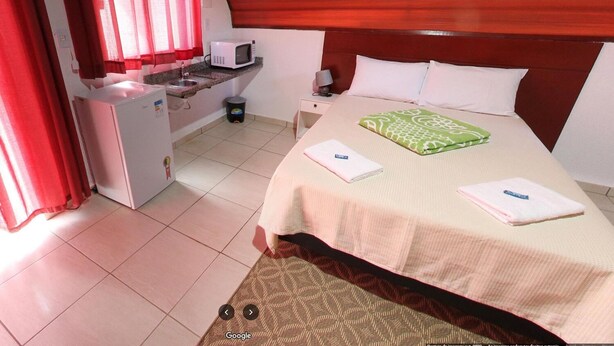 A hotel room with a bed, a desk with a microwave, a small trash bin, and a window with red curtains. - Espaço Chalé Cereja (Cachoeira Paulista)