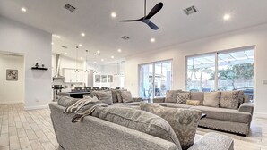 A spacious living room with a large grey sofa set, a dining area with a kitchen in the background, and large windows providing natural light.