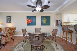 A dining area with a glass table, wicker chairs, a ceiling fan, and artwork on the walls.