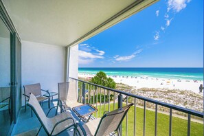 Un balcon avec des chaises et une table, donnant sur une plage parsemée de parasols et fréquentée par des gens.