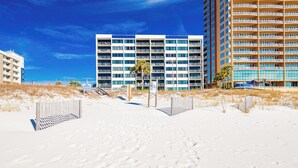 Un hotel frente a la playa con varios balcones, una playa arenosa en primer plano y un cielo azul despejado.