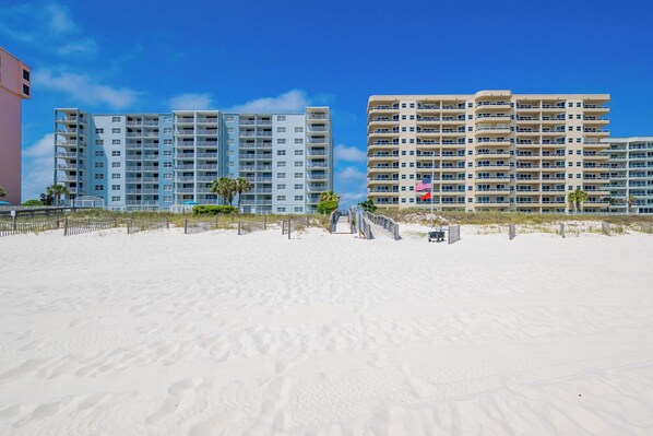 Un hotel frente a la playa con varios pisos, una playa arenosa en primer plano y un cielo azul despejado.