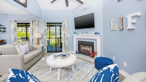 A living room with a fireplace, a ceiling fan, a flat-screen TV, and a large window with curtains. - Beautifully Decorated HOME! ~ Close to EVERYTHING! ~ 2BR 2BA ~ Mariner Lakes (Orange Beach)