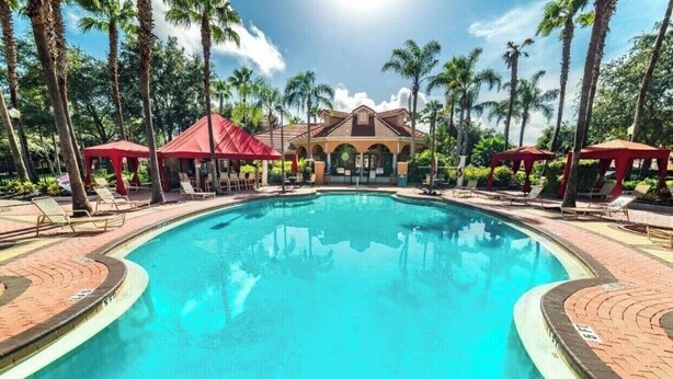 Una piscina con sillas de descanso y toldos rojos, rodeada de palmeras y un edificio con techo de tejas.