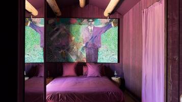 A bedroom with a wooden ceiling, a large abstract painting, a bed with red bedding, and a purple ottoman.