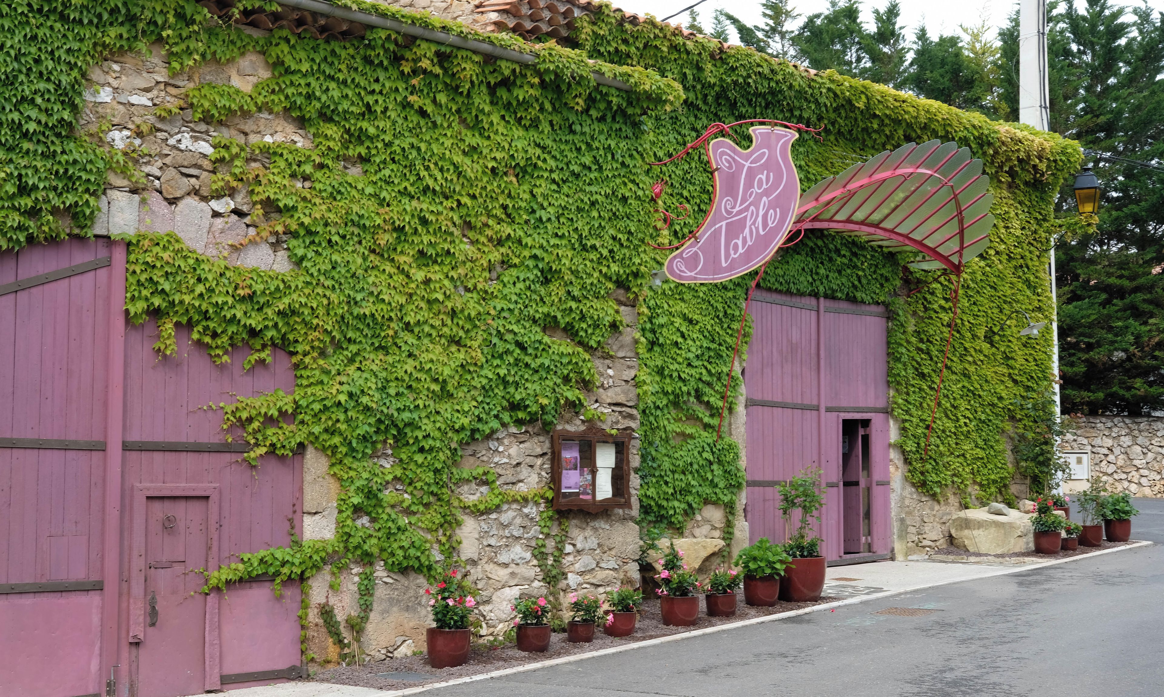 Un edificio con pareti ricoperte d'edera, un tendone viola e un cartello che recita 'La Table'.