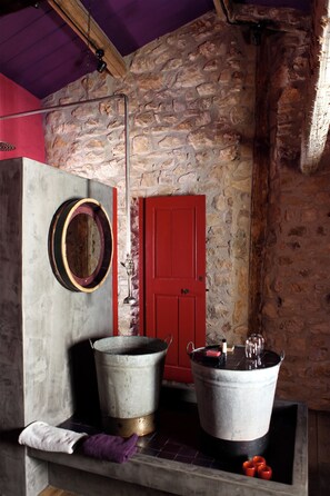 Un interior rústico con una pared de piedra, una puerta roja, un balde de metal y un espejo colgado.