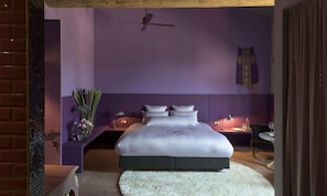 A bedroom with a large bed, a ceiling fan, a nightstand with a lamp, and a decorative wall hanging.