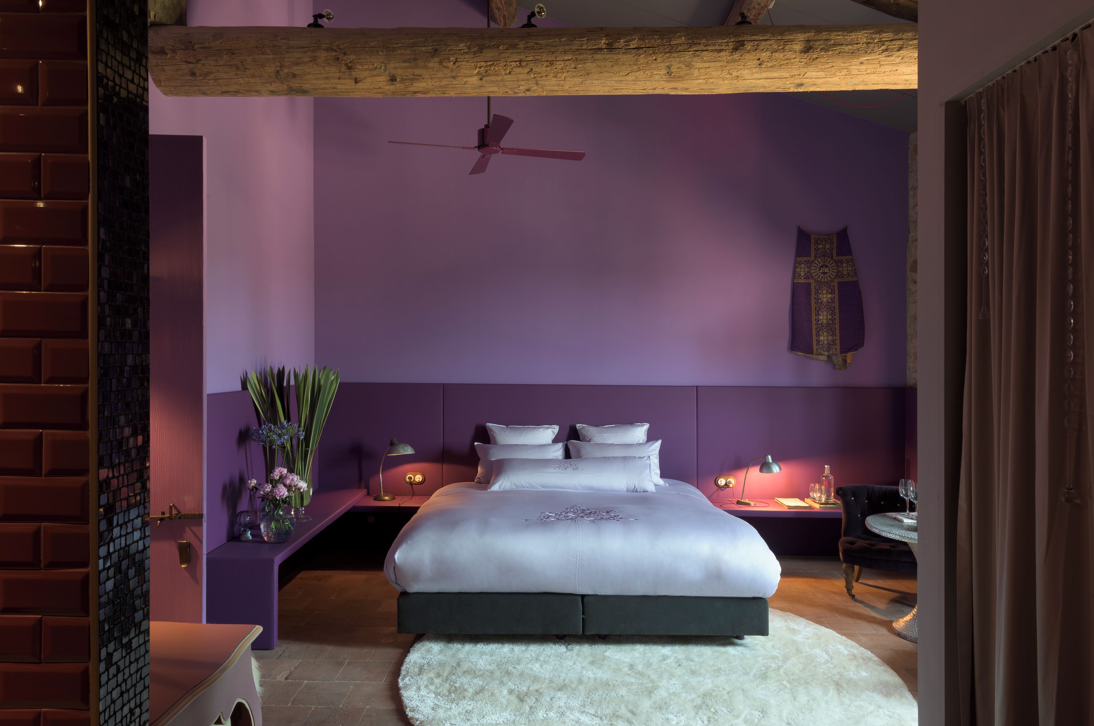 A bedroom with a large bed, a ceiling fan, a cross on the wall, and a nightstand with a lamp.