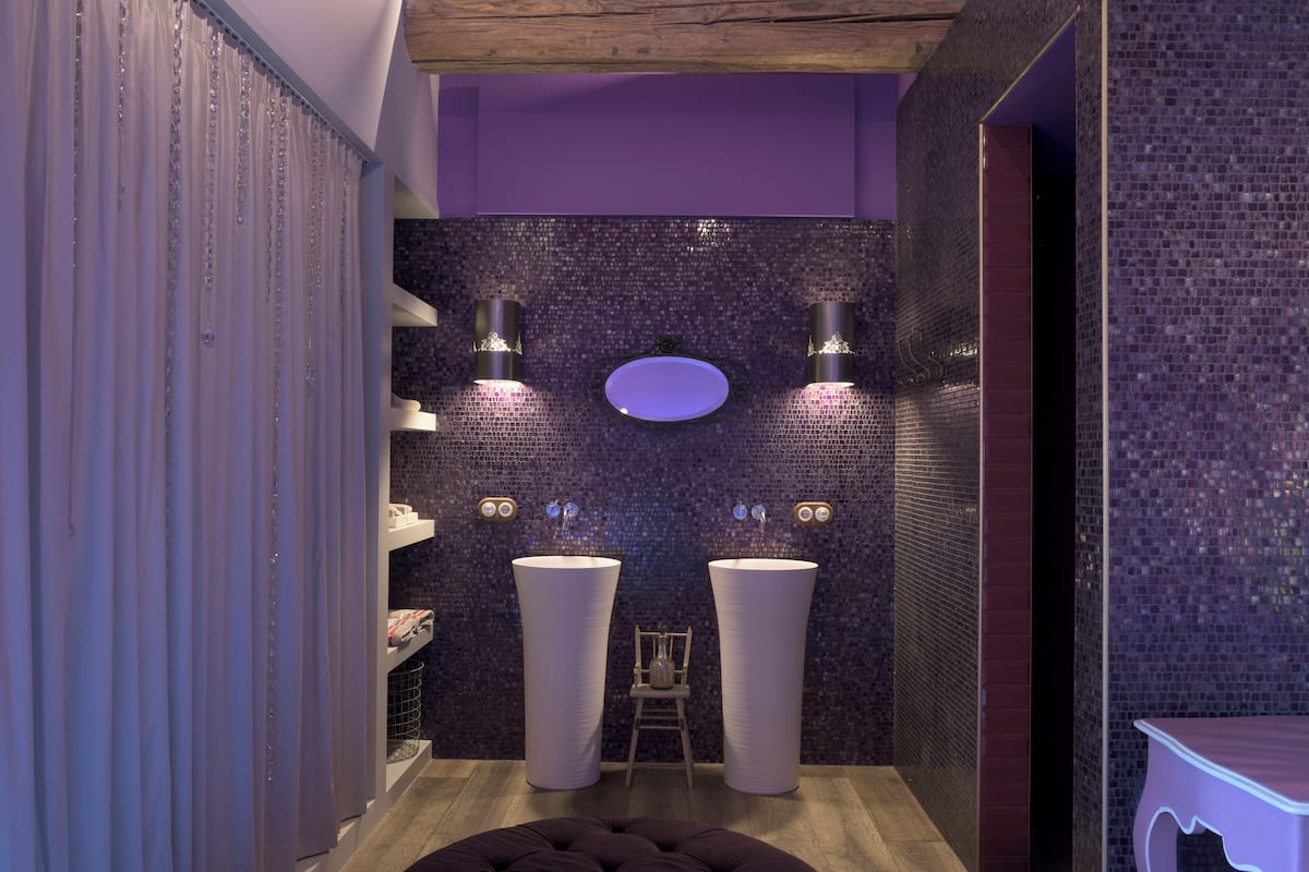 A modern bathroom with two large sinks, a small wooden chair, and a purple tiled wall.