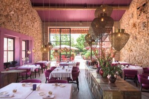 A restaurant with stone walls, purple accents, and hanging light fixtures. Tables are set with white tablecloths and pink chairs.